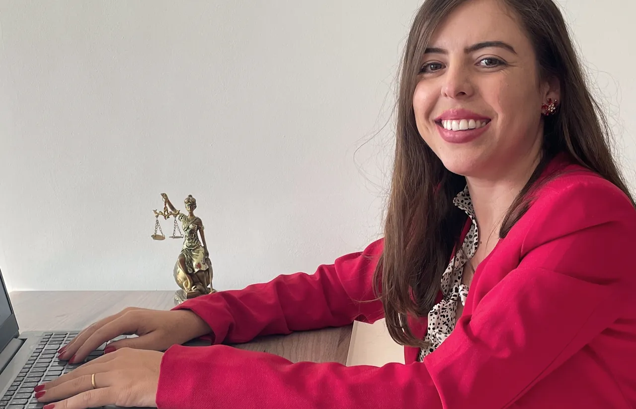 Dra. Fernanda Márcia Ferreira Guedes, de blazer vermelho, sorrindo em sua mesa de atendimento, com a estátua da Justiça ao fundo